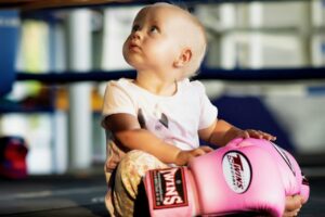 Adorable baby girl sitting on the floor with pink boxing gloves, symbolizing strength and gentle postpartum recovery